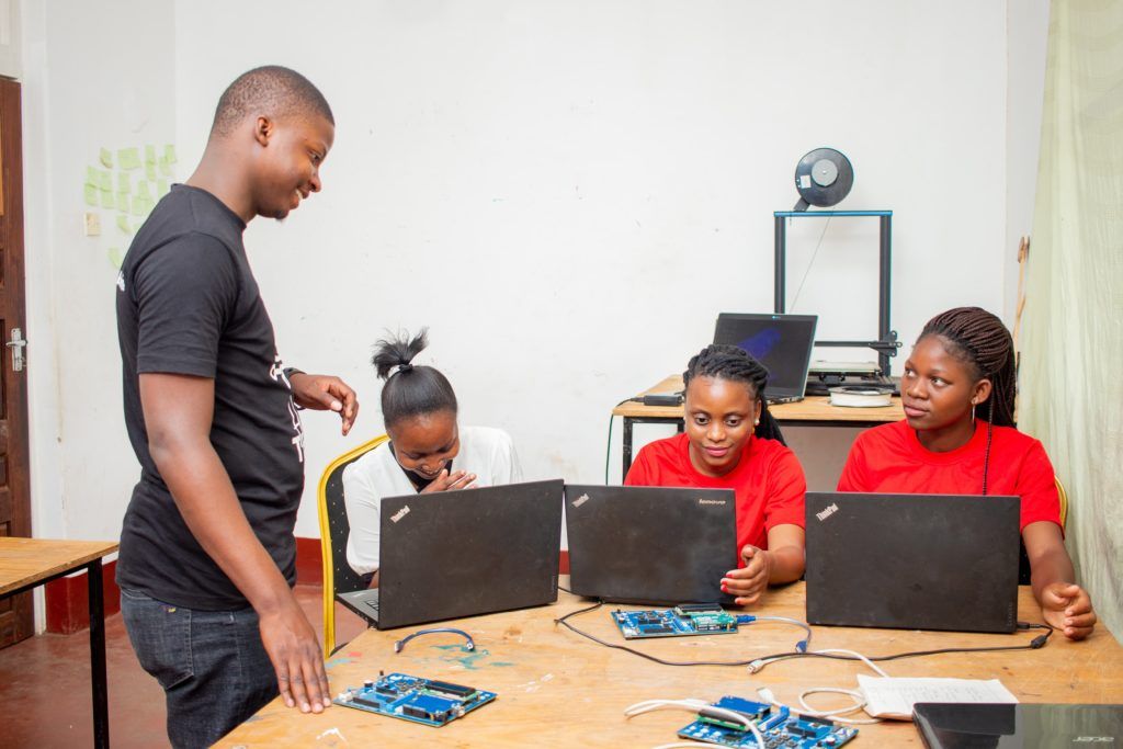 three happy, young African women sitting at a desk in front of their laptops and listening to an young African man standing next to the desk